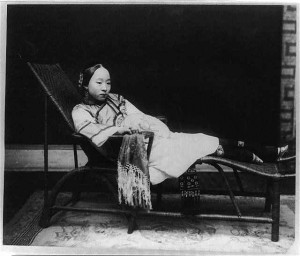 "This text represents a few of the most pervasive motivations and ideas associated with footbinding in the nineteenth and twentieth century. It captures the essence of social structure in China at a time when male dominance and masculinity relied heavily on female footbinding. In addition, this photo symbolizes many valued aspects of footbound women at the time; concealment, mysteriousness, elegance, and luxury.   Upon seeing this photo, the viewer may question why the woman is sitting in a chair. Although her feet are not a dominant feature in this image, we are able to see they have been bound. Many women in the twentieth century were forced to work physically demanding jobs with crippled feet, but footbinding as fashion culture disabled many women from certain tasks completely. This female weakness and sense of helplessness gave males a chance to further assert their masculinity and power. The woman’s bound feet are not the prominently displayed in this text to represent the mysteriousness of footbinding culture. The photographer uses darkness around the bound feet strategically to proclaim this mysteriousness. In addition, intricately embroidered shoes cover her feet giving the woman the ability to conceal them. Women with bound feet were respected and their decision to participate in the fashion system displayed their civility and political obedience. Rather than placing her feet on the floor, the photographer chooses to keep them elevated to display their value and elegance. Finally, this woman dressed in silk, rests on a chaise, symbolizing luxury and high class."