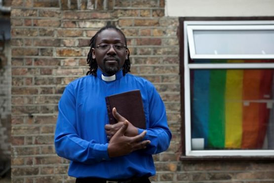 Preacher holding a Bible with a Pride Flag hanging from a window in the background.