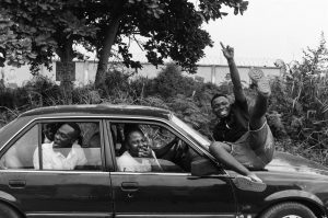 Five men (four in a car and one on top of the hood) smiling and having a good time
