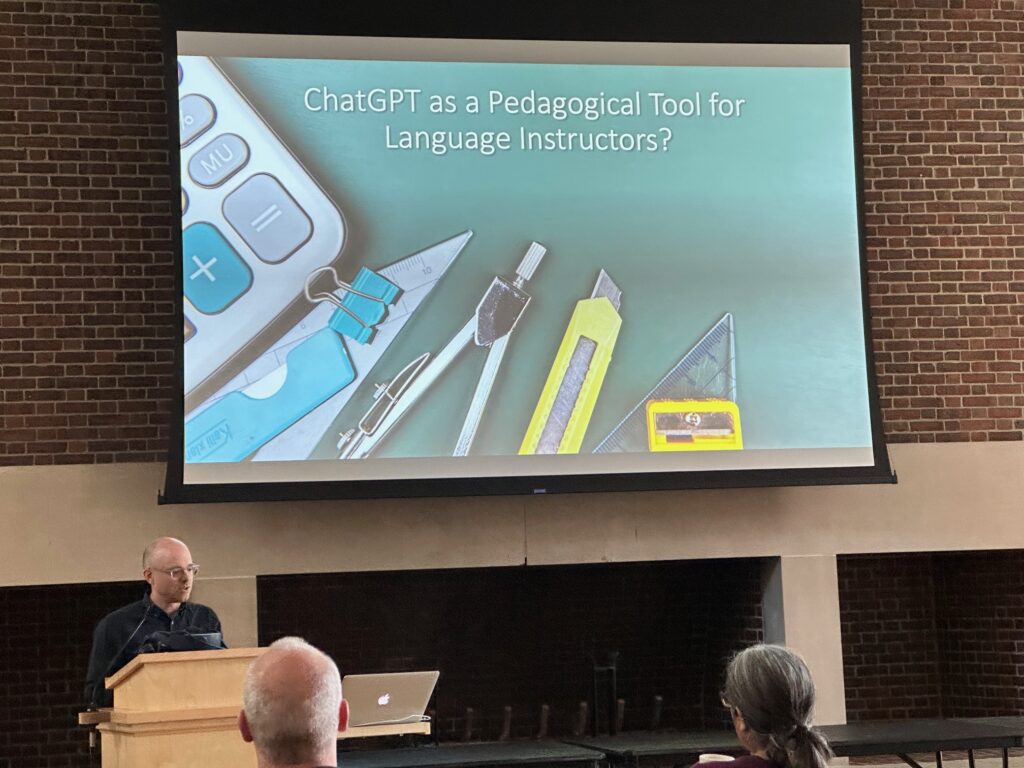 Professor Reed Johnson standing at a podium with a PowerPoint slide showing on a screen behind him that says ChatGPT as a Pedagogical Tool for Language Instructors.