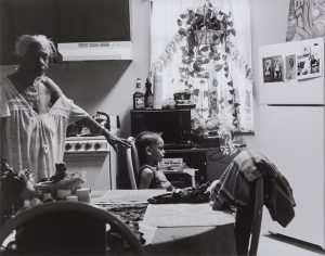 Photograph of an elderly Black woman standing and resting her proper left arm against a chair where a young Black boy is seated. Both people are in a kitchen