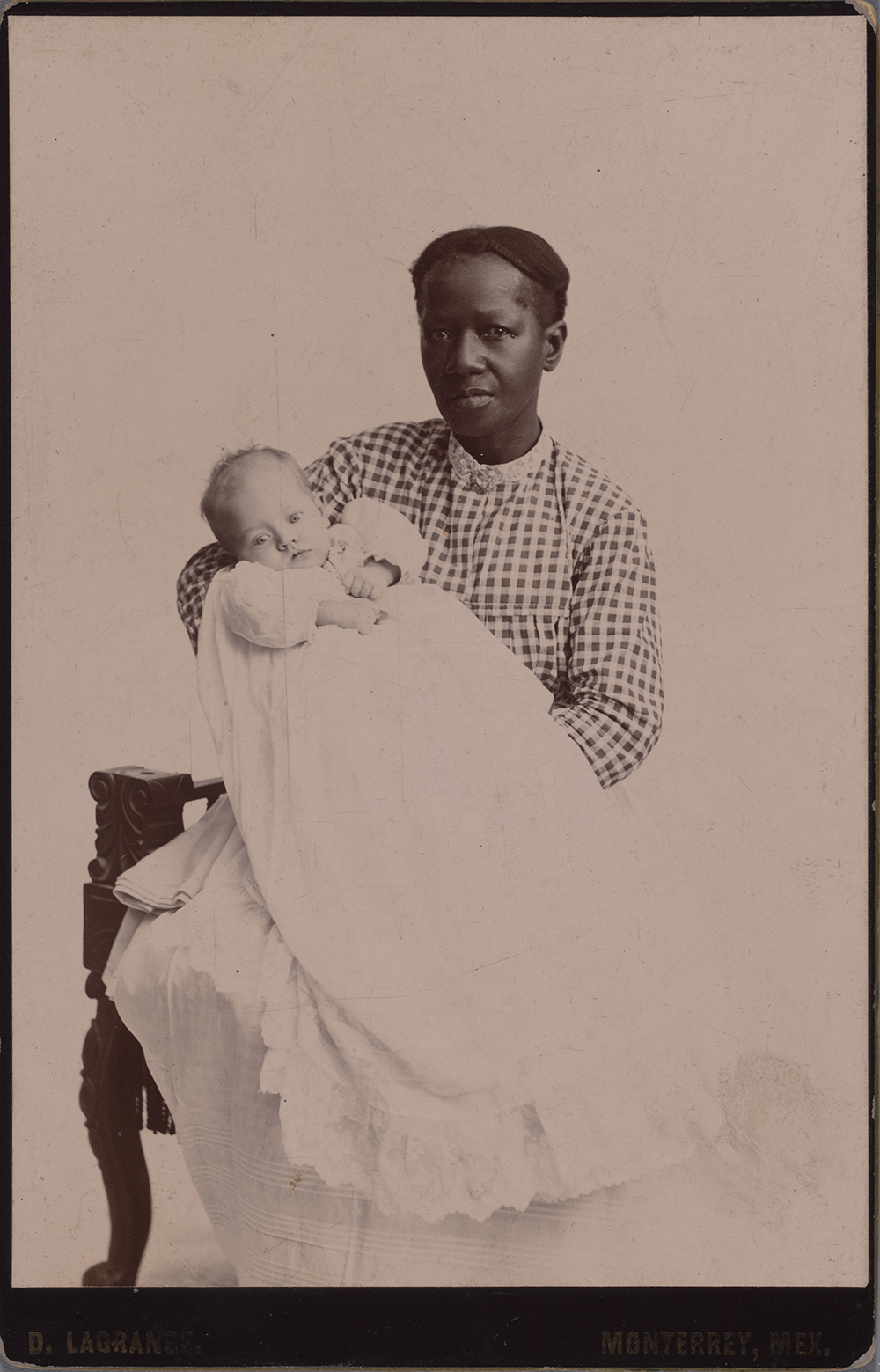 Photograph of a Wet Nurse of African Descent with braided hair and a checkered shirt holding a white infant in a christening gown