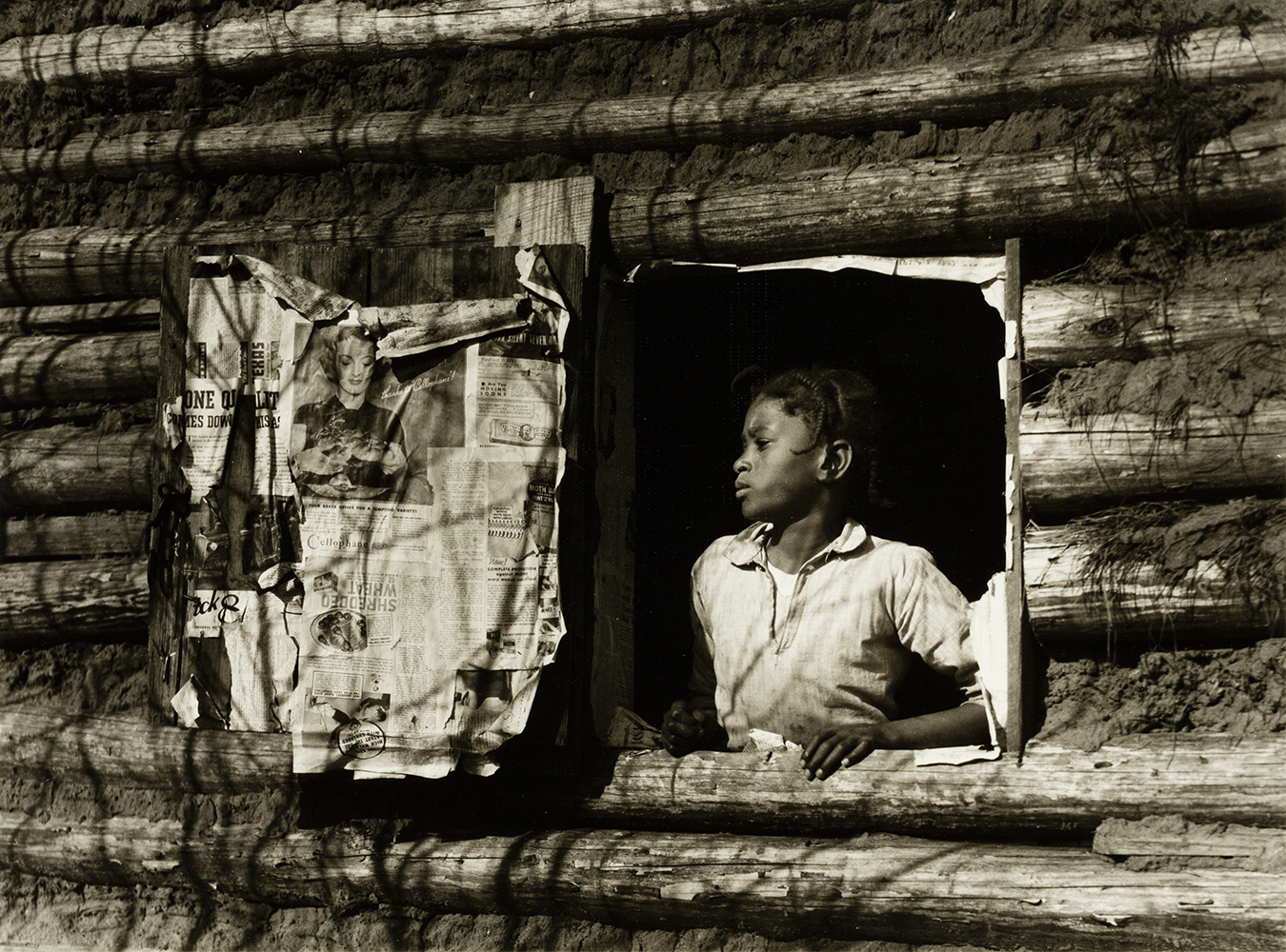 Photograph of a young Black girl staring out of an open window of a log home. The interior of the window door and home contain layers of newspaper on the walls