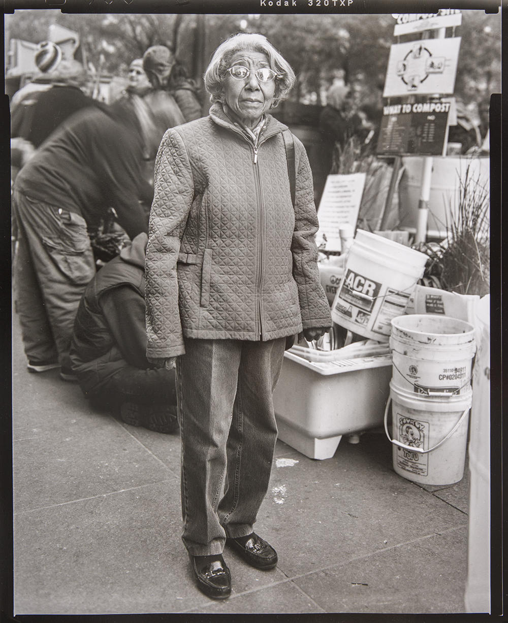 Photograph of an elderly Black woman in a quilted jacket standing outside on a busy street in Manhattan during the Occupy Wall Street protest