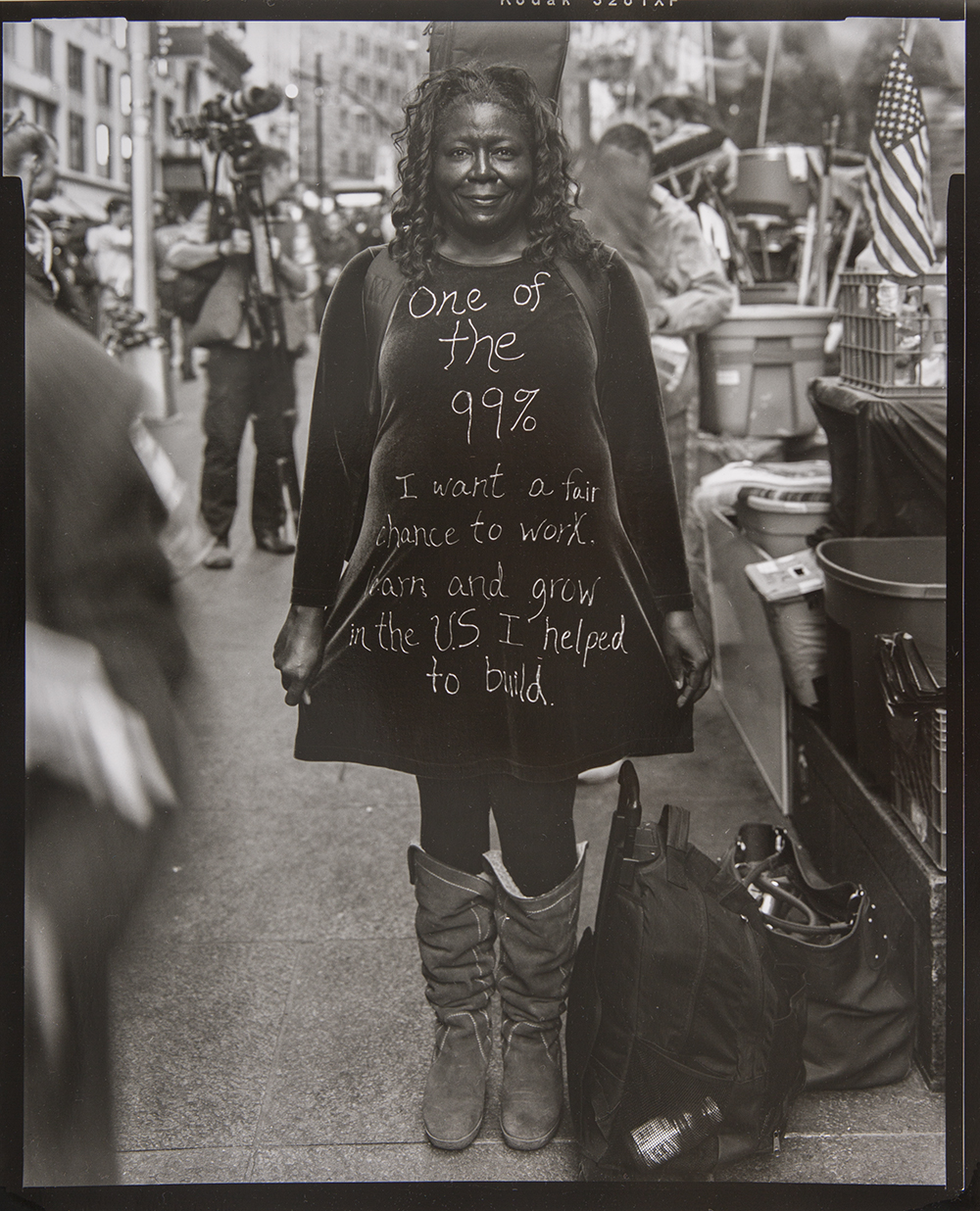 Photograph of a Black woman standing outside on a busy street in Manhattan during the Occupy Wall Street protest wearing a dress that reads: “One of the 99%. I want a fair chance to work, learn and grow in the U.S. I helped to build.”