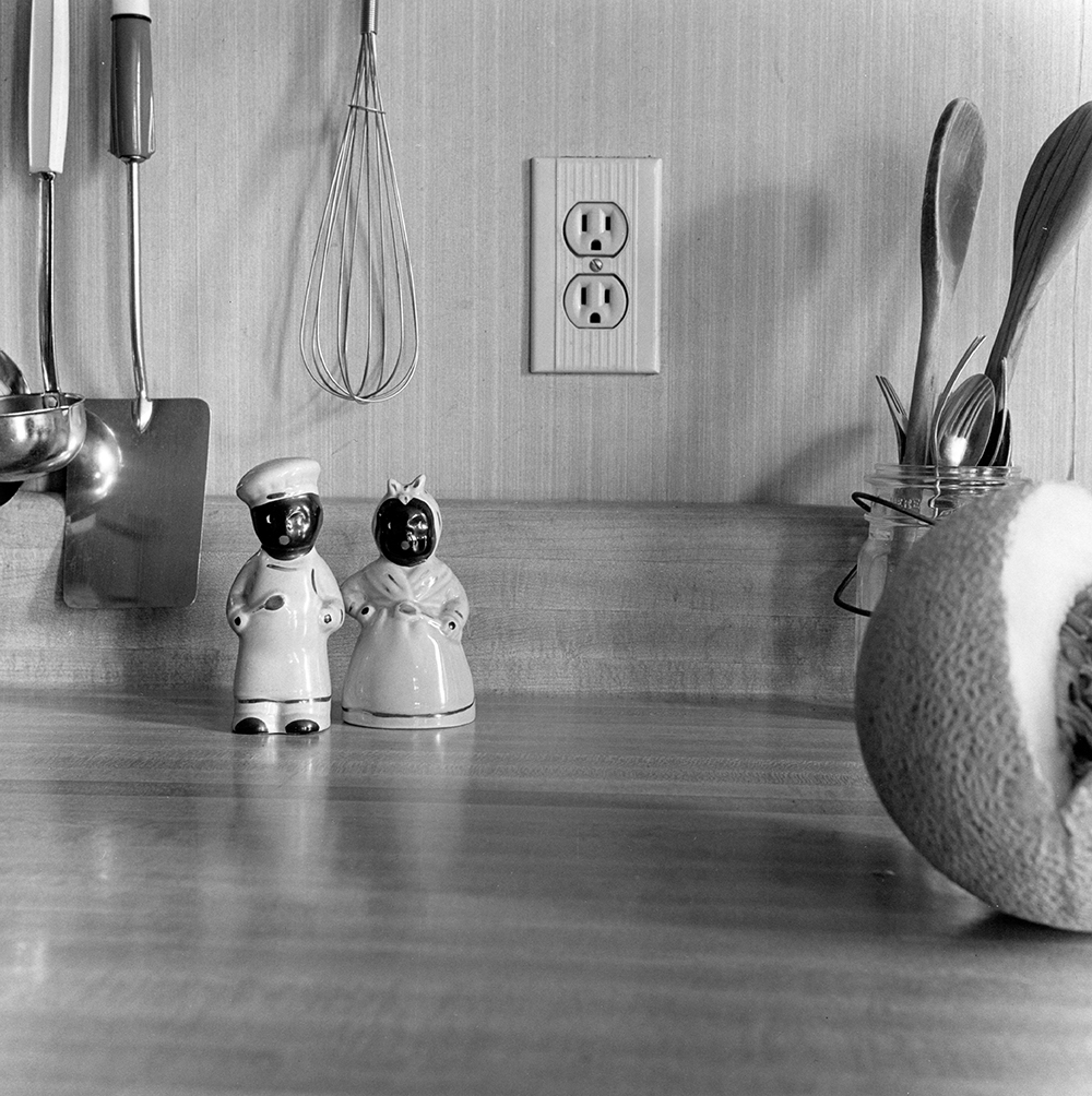 Photograph of a salt and pepper shaker set depicting Sambo and Mammy stereotypes sitting on a domestic kitchen counter with silverware, utensils, and a cut cantaloupe