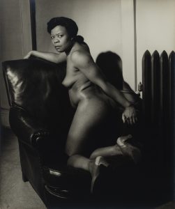 Photograph of a nude Black woman kneeling in a large leather chair, head turned towards the viewer