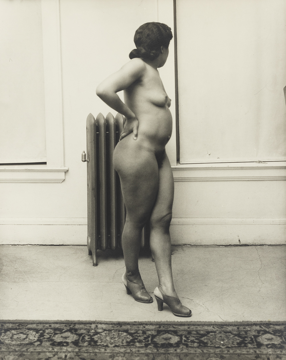 Photograph of a nude Black woman turned in profile standing in a sparse room in front of a hot water radiator
