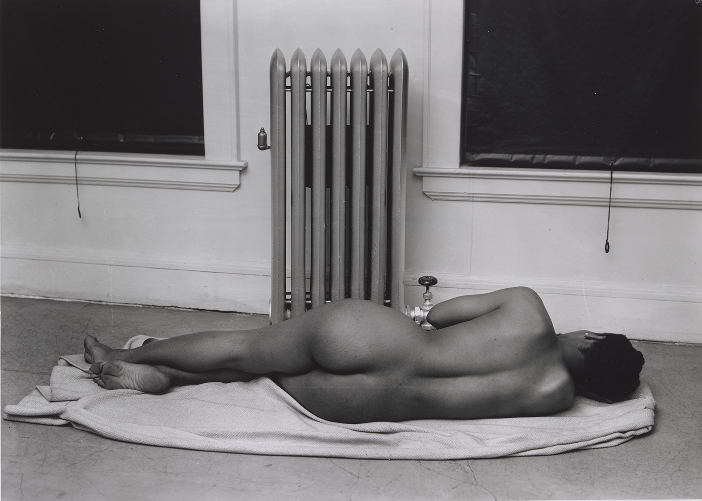 Photograph of a nude Black woman laying down on her side, back towards the viewer, in a sparse room in front of a hot water radiator