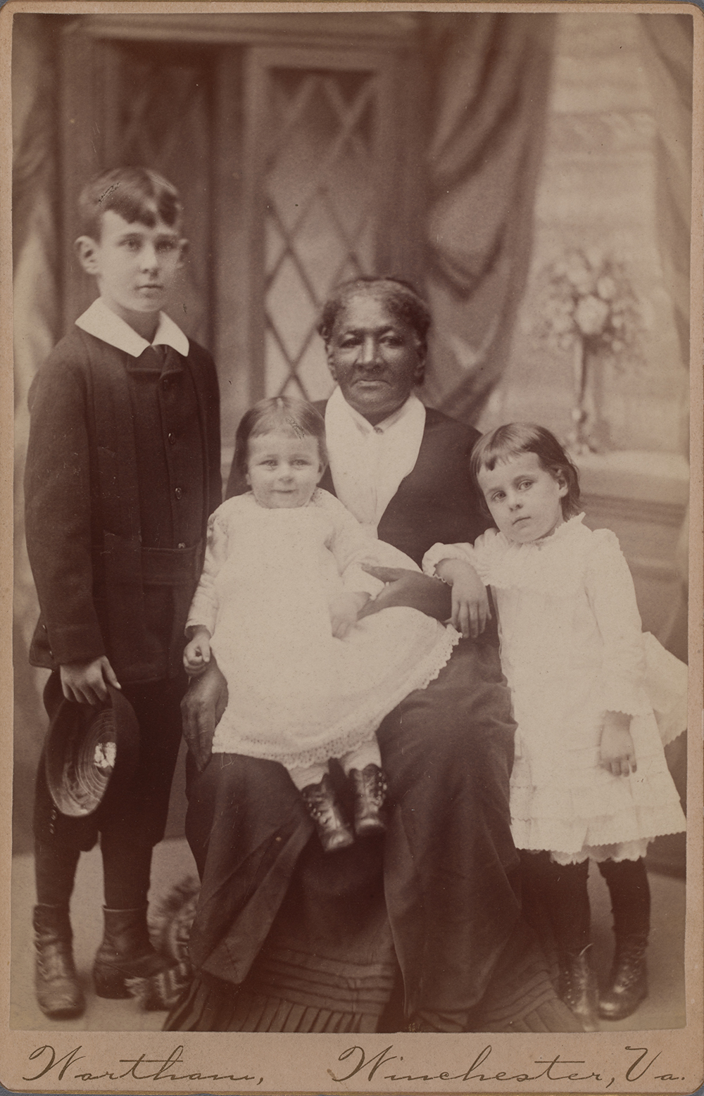 Photograph of an elderly Black woman seated surrounded by a white infant seated on the woman’s proper right knee, a young white girl resting against the woman at her proper left, and a young white boy standing at the woman’s proper right. All subjects are against a studio photography backdrop