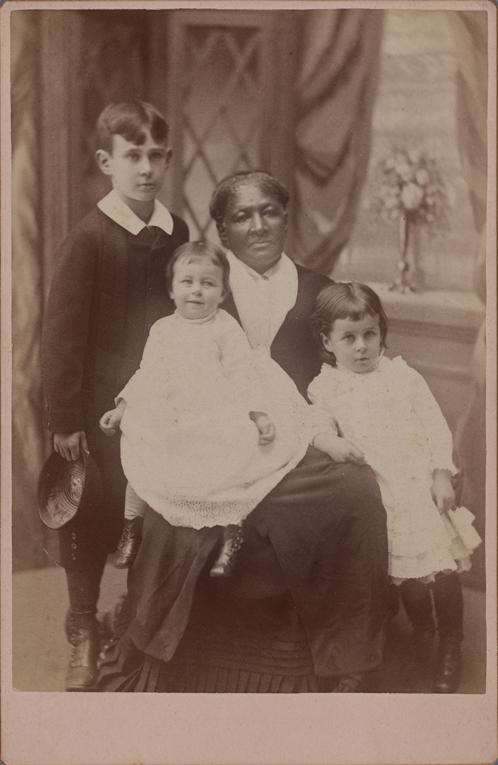 Photograph of an elderly Black woman seated surrounded by a white infant seated on the woman’s proper right knee, a young white girl resting against the woman at her proper left, and a young white boy standing at the woman’s proper right. All subjects are against a studio photography backdrop